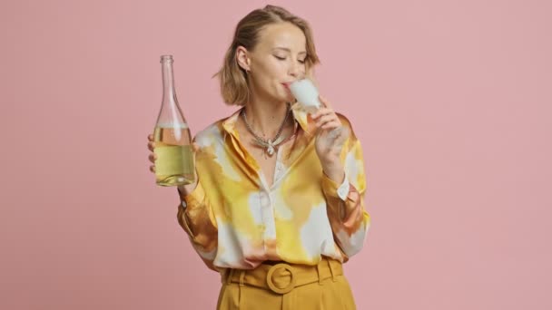 幸せな可愛いですブロンド女性でカラフルなエレガントな衣装飲みシャンパンと見ますザカメラ上のピンクの背景 — ストック動画
