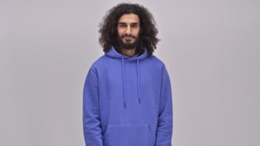 Uzun kıvırcık saçlı yakışıklı, sakallı esmer bir adam stüdyodaki gri duvarın üzerinden kameraya bir öpücük yolluyor.