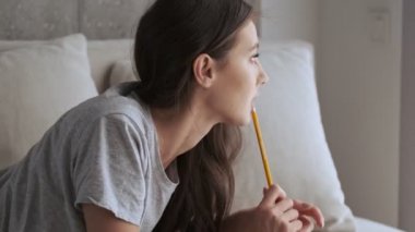 Evdeki kanepede yatarken elinde kalem tutan ve başka tarafa bakan düşünceli, güzel esmer kadın.