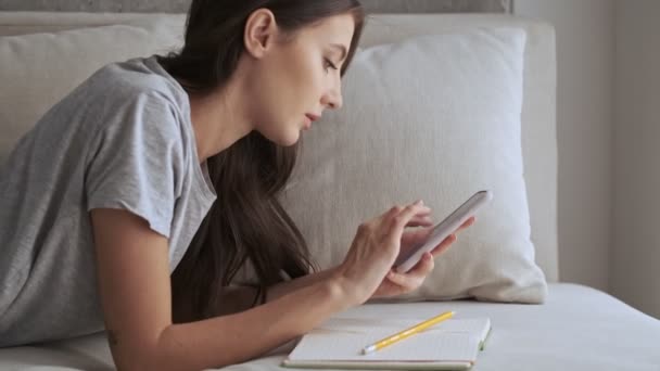 自宅でソファに横たわっている間 スマートフォンを使用してノートブックに何かを書く穏やかなかなりブルネットの女性の側面図 — ストック動画