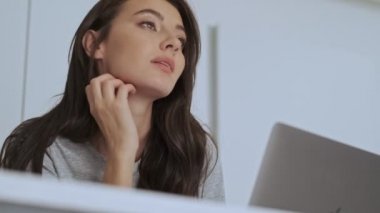 Düşünceli, güzel esmer bir kadın evde masada otururken gözlerini kaçırıp dizüstü bilgisayar kullanıyor.
