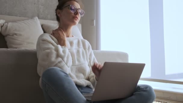 幸せなかわいいですブルネット女性で眼鏡見ながら離れて座っている間に窓の近くにラップトップコンピュータとともに自宅 — ストック動画
