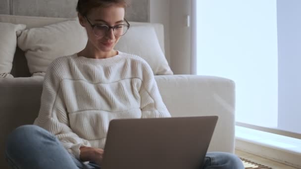 家の窓の近くに座っている間に ラップトップコンピュータを使用して眼鏡の中でかなりブルネットの女性を落ち着かせ 疲れになる — ストック動画