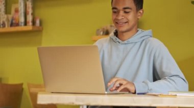 Pozitif neşeli, genç Afrikalı öğrenci ödevlerini yaparken dizüstü bilgisayar kullanıyor..