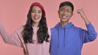 Genç, mutlu, sevgi dolu bir çift pembe duvar arkasında izole bir şekilde baş parmaklarını gösteriyor.