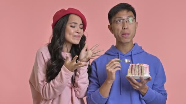 年轻饥饿的女人看着她的男朋友是如何在粉红墙的背景下吃一个孤立的蛋糕的 — 图库视频影像