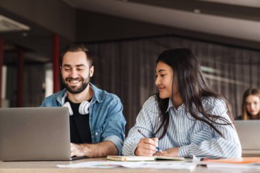 Çok uluslu neşeli öğrencilerin sınıfta çalışırken dizüstü bilgisayar kullandıkları ve egzersiz kitabına yazdıkları fotoğraf.
