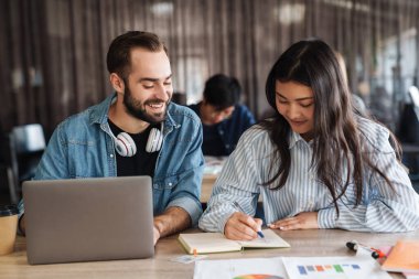 Çok uluslu neşeli öğrencilerin sınıfta çalışırken dizüstü bilgisayar kullandıkları ve egzersiz kitabına yazdıkları fotoğraf.