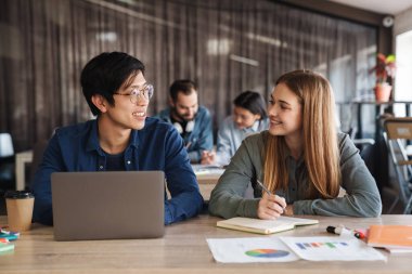 Çok uluslu neşeli öğrencilerin sınıfta çalışırken dizüstü bilgisayar kullandıkları ve egzersiz kitabına yazdıkları fotoğraf.