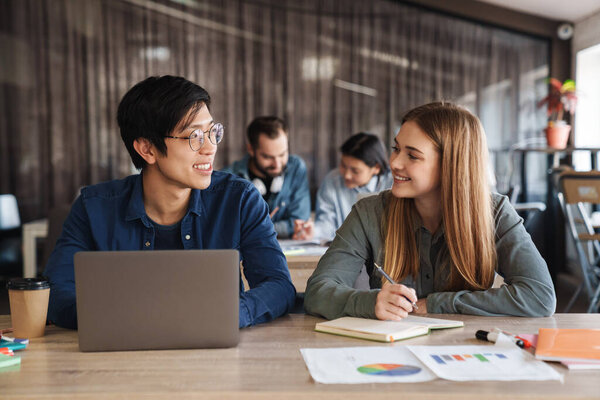 Photo of multinational joyful students using laptop and writing in exercise book while studying at classroom