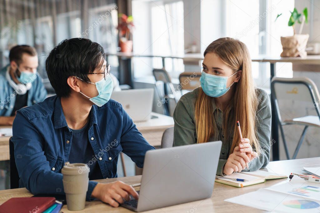 Image of multinational focused students in protective mask talking while studying with laptop at classroom
