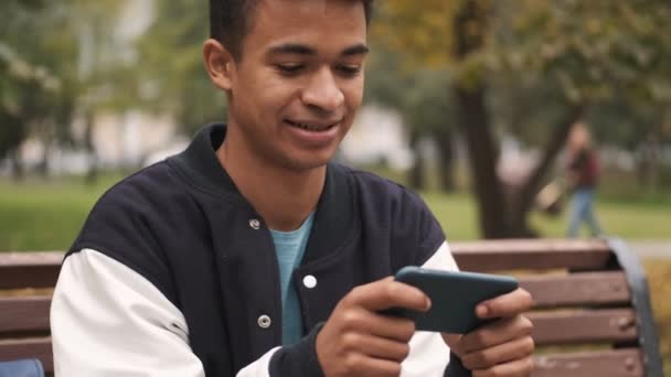 一个情绪激动的非洲小伙子学生坐在长椅上 用手机在室外玩游戏 — 图库视频影像