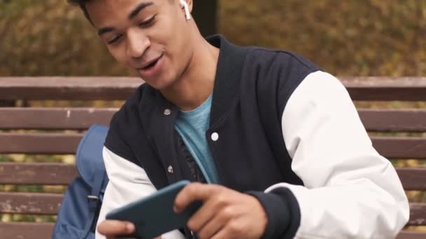 Geconcentreerde Jonge Emotionele Afrikaanse Jongen Student Oortelefoons Zitten Bank Spelen — Stockvideo