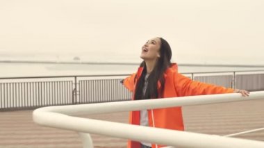 Güzel Asyalı neşeli pozitif kadın limandan geçiyor açık hava bursu turuncu ceketli ve elleri havada..
