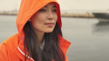 Güzel Asyalı memnun kadın limandan geçiyor açık havada turuncu ceketli kablosuz kulaklıklarla müzik dinliyor..