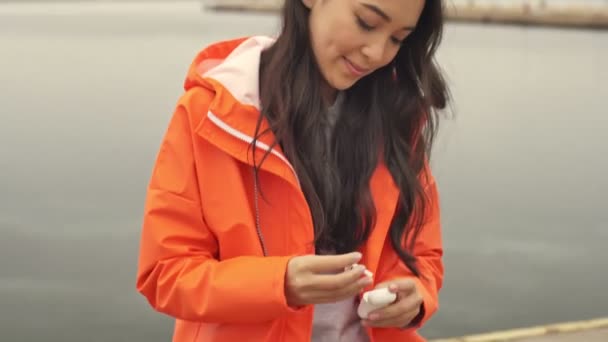 驚くべきアジアの幸せな陽気な女性歩くことによって港助成金屋外でオレンジコートリスニング音楽で無線イヤフォン — ストック動画