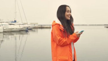 Çekici Asyalı neşeli kadın liman tarafından yürüyor turuncu ceketli açık hava bursu cep telefonu sohbetini kullanarak.