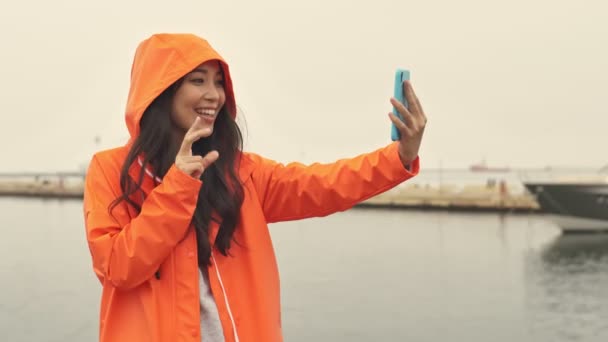 漂亮的亚洲女人穿着橙色外套走在港口外的赠品旁 用手机自拍 — 图库视频影像
