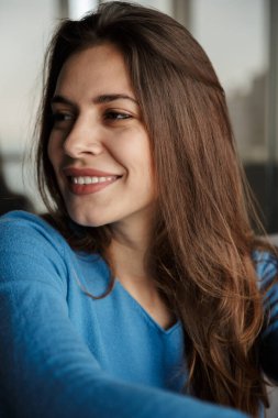 Oturma odasında kanepede oturmuş gülümseyen neşeli genç bir kadın görüntüsü.