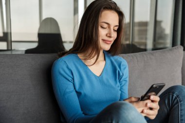 Kafkasyalı neşeli genç bir kadının oturma odasındaki kanepede otururken cep telefonu kullanması ve gülümsemesi.