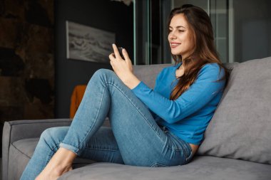 Oturma odasındaki kanepede otururken cep telefonu kullanan ve gülümseyen güzel bir genç kadın görüntüsü.