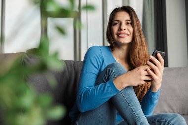 Oturma odasındaki kanepede otururken cep telefonu kullanan ve gülümseyen güzel bir genç kadın görüntüsü.