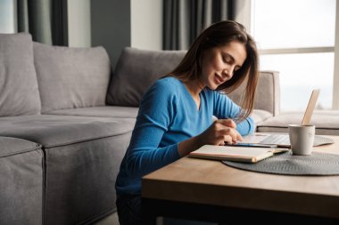 Oturma odasında yerde otururken dizüstü bilgisayar ve cep telefonu kullanan gülümseyen genç bir kadın görüntüsü