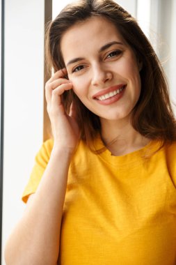 Memnun genç ve güzel bir kadının, pencerenin yanında durup kameraya bakıp gülümsemesi.