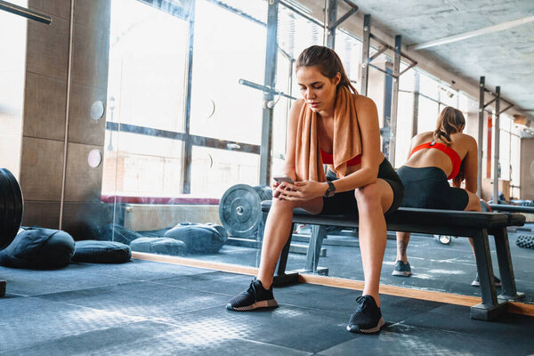 Attractive young fit sportswoman resting after exercises while sitting in the gym, using mobile phone