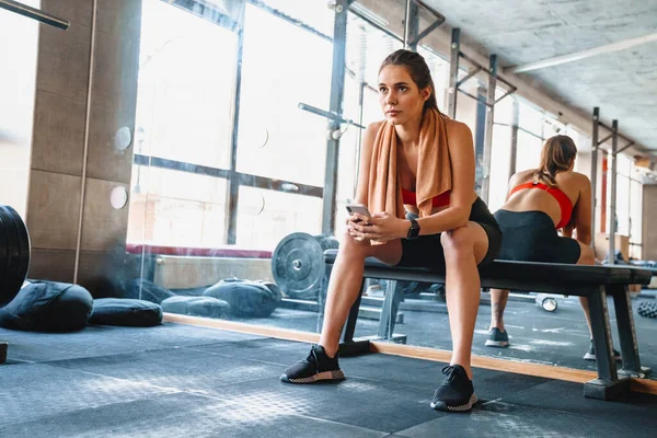 有魅力的年轻健康的女运动员坐在体育馆里 一边锻炼 一边用手机休息 — 图库照片