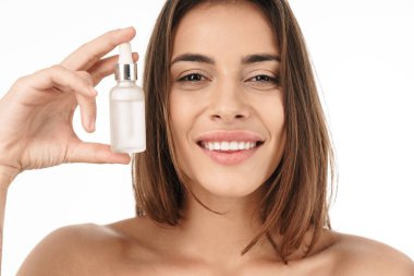 Çekici, gülümseyen, üstsüz bir kadının portresi beyaz arka planda izole bir şekilde duruyor, yüz yağı şişesini gösteriyor.