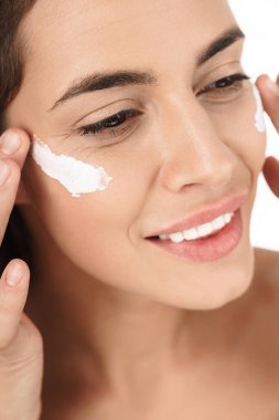 Beyaz arka planda izole edilmiş, gülümseyen ve yüz kremi süren pozitif, yarı çıplak bir kadın portresi.