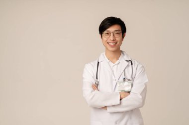 Tıbbi beyaz üniformalı Asyalı genç doktor bej arka planda izole bir şekilde gülümsüyor.