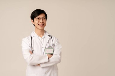 Tıbbi beyaz üniformalı Asyalı genç doktor bej arka planda izole bir şekilde gülümsüyor.