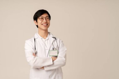 Tıbbi beyaz üniformalı Asyalı genç doktor bej arka planda izole bir şekilde gülümsüyor.