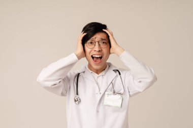 Beyaz üniformalı Asyalı genç doktor bej arka planda izole edilmiş paniği haykırıyor ve ifade ediyor.