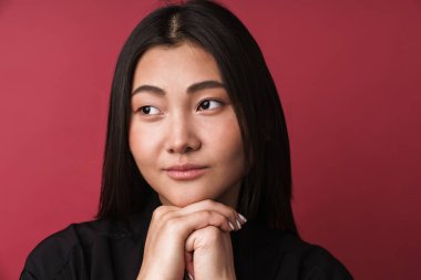 Oldukça düşünceli, genç Asyalı bir kadının kırmızı arka planda tek başına dikilip bakışlarını kaçırmasını.