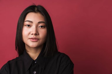 Kendine güvenen, sıradan kıyafetler giyen, kırmızı arka planda tek başına duran, kameraya bakan Asyalı genç bir kadının yakın çekimi.