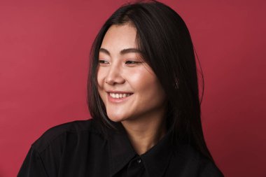 Kırmızı arka planda tek başına duran, sıradan kıyafetler giyen, gülümseyen genç Asyalı bir kadının fotoğrafını çek.