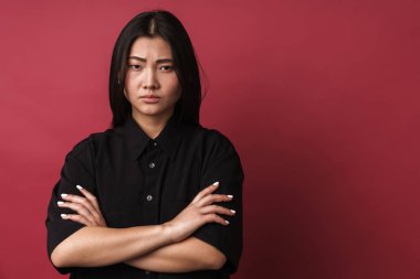 Kırmızı arka planda tek başına duran, sıradan kıyafetler giyen oldukça üzgün Asyalı genç bir kadının yakın plan fotoğrafı.