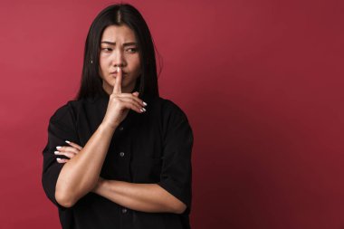 Kırmızı duvar arkasında izole edilmiş güzel bir Asyalı genç kadının görüntüsü sessizlik hareketi gösteriyor..