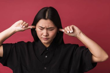 Kızıl duvar arkasında izole edilmiş kulakları kaplayan oldukça hoşnutsuz Asyalı genç bir kadının görüntüsü.