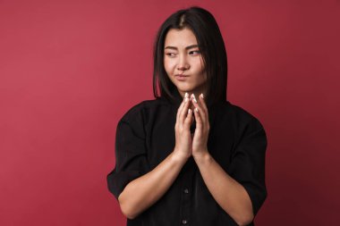 Kırmızı arka planda tek başına duran, sıradan kıyafetler giyen oldukça üzgün Asyalı genç bir kadının yakın plan fotoğrafı.