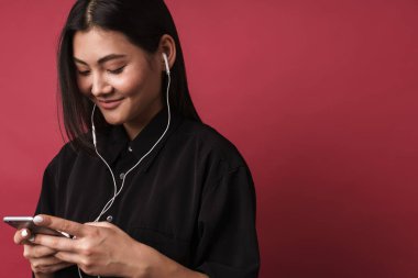 Kırmızı arka planda izole bir şekilde dikilmiş, kulaklıkla müzik dinleyen, elinde akıllı telefon tutan, güzel gülümseyen Asyalı genç bir kadının fotoğrafını çek.