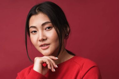 Kendine güvenen, sıradan kıyafetler giyen, kırmızı arka planda tek başına duran Asyalı genç bir kadına yakın durun.