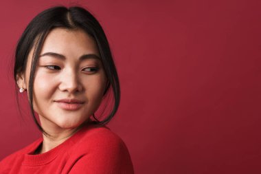 Kırmızı arka planda tek başına duran, sıradan kıyafetler giyen, gülümseyen genç Asyalı bir kadının fotoğrafını çek.