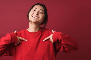 Kırmızı arka planda tek başına kendini işaret eden, sıradan kıyafetler giyen, gülümseyen, genç Asyalı bir kadını yakından çek.