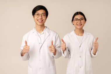 Beyaz üniformalı Asyalı genç tıp doktorlarının bej arka planda izole edilmiş gülümseyen ve işaret eden başparmakları.