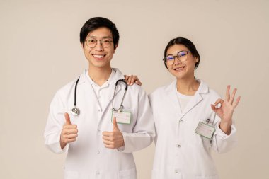 Beyaz üniformalı Asyalı genç tıp doktorlarının bej arka planda izole edilmiş, gülümseyen ve iyi jestler yapmaları.