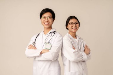 Beyaz üniformalı Asyalı genç tıp doktorlarının bej arka planda izole edilmiş bir şekilde ayakta durup gülümsemesi.